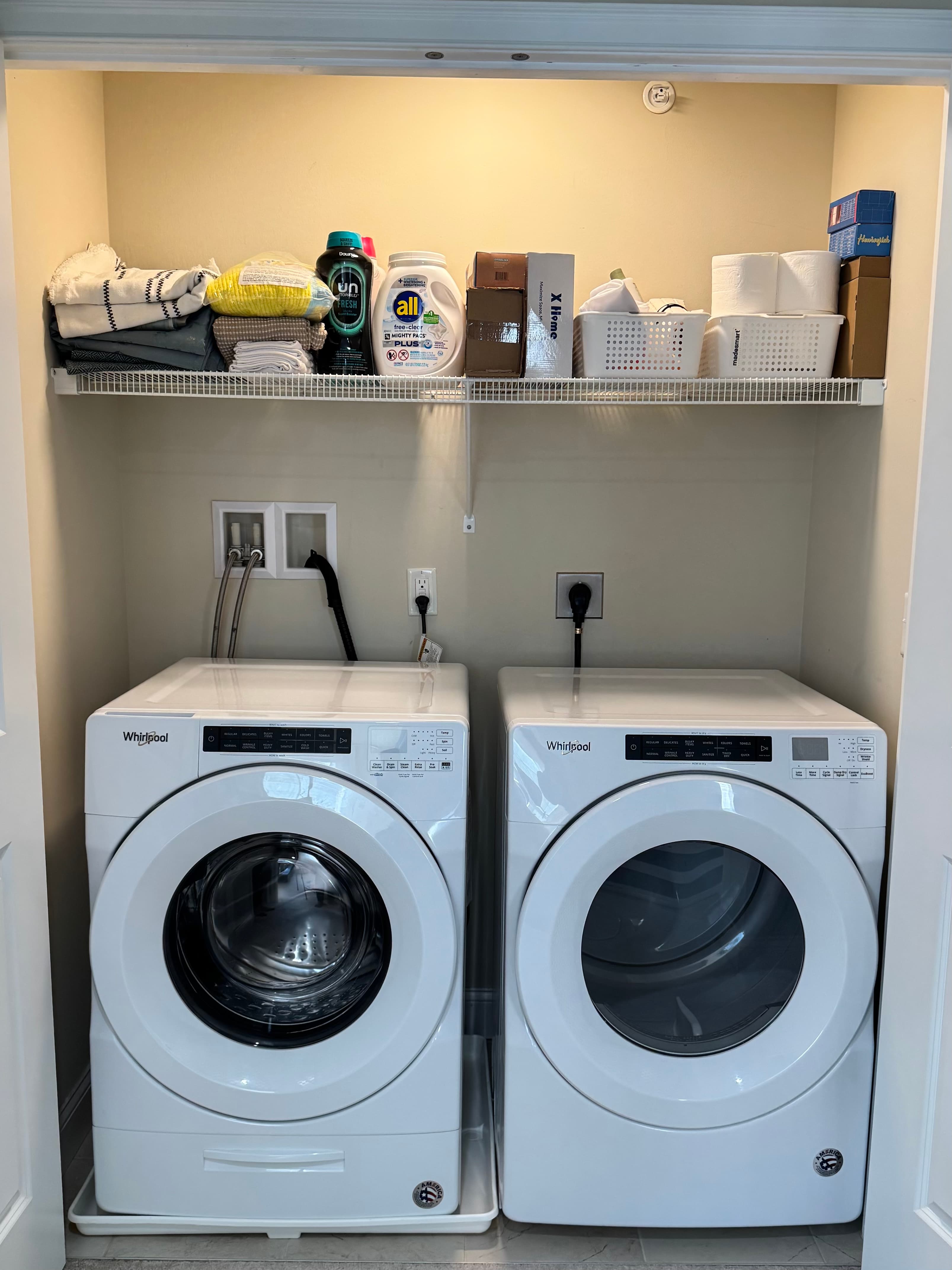 Wire shelf above a laundry closet holding detergent bottles, a cardboard box, and stacked paper towels before organizing